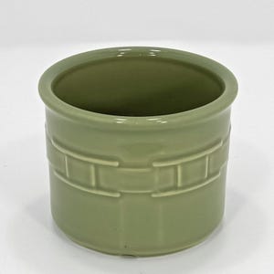 May include: A sage green ceramic container with a cylindrical shape. The container has a decorative band of rectangular indentations around the middle. The interior is smooth and the rim is slightly flared.