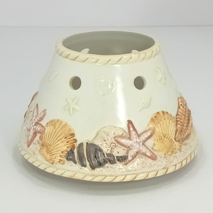 May include: Cream-colored ceramic candle warmer with a beach theme. The warmer features embossed seashells, starfish, and sand dollar designs. It has two holes for ventilation and a rope-like trim around the top and base.