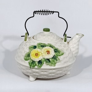 May include: White ceramic teapot with a woven basket design and floral accents. The teapot has a black wire handle with a spring detail and a lid with a green knob.