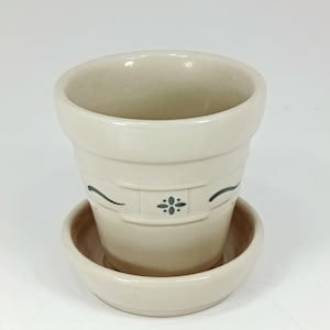 May include: A cream-colored ceramic flower pot with a matching saucer. The pot has a slightly textured surface and is decorated with a dark green floral design. The pot is cylindrical with a flared top.