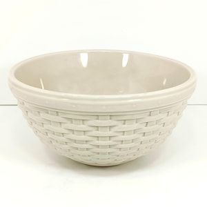 May include: A cream-colored ceramic bowl with a woven basket design. The bowl has a smooth, rounded interior and a textured exterior that mimics a basket weave. The rim features a decorative beaded detail.