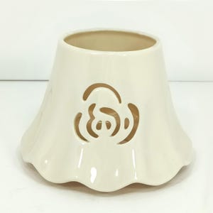 May include: Off-white ceramic candle holder with a flared, ruffled base. The body features a cut-out rose design. The interior is a lighter shade of off-white. The candle holder is designed to hold a small candle.
