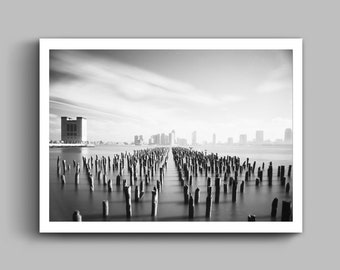 May include: A black and white photograph of a city skyline with a long row of wooden pilings in the foreground. The pilings are partially submerged in the water and create a sense of depth and perspective. The city skyline is in the distance and is slightly blurred, creating a sense of mystery and intrigue.