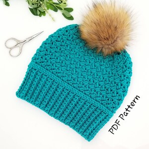 Op de afbeelding: Turquoise blauwe gehaakte beanie met een bruine nep bont pompon. De beanie heeft een geribbelde rand en een textuur steekpatroon. PDF-patroon.