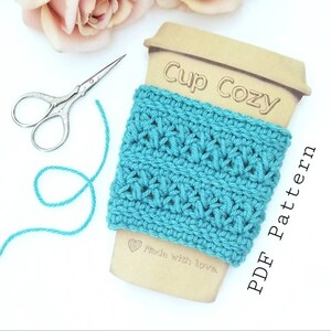 May include: A teal blue crocheted cup cozy with the text "Cup Cozy" and "PDF Pattern" on a wooden background. The text "Made with love." is also on the background.