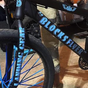 May include: A matte black bicycle featuring bright blue spokes and light blue decals. The frame displays the text "BLOCKS FLYER" and "LANDING GEAR" on the front fork. The bike includes black pedals, a black seat, and a black chainring assembly.