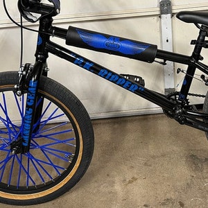Könnte beinhalten: Schwarzes BMX-Fahrrad mit blauen Akzenten. Das Fahrrad hat blaue Speichen, einen Lenker und einen Rahmenschutz mit blauem Design. Die Reifen haben eine beige Seitenwand. Der Text "SE RACING" ist auf dem Rahmen zu sehen.