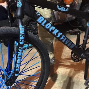May include: Black bicycle with blue accents, including the spokes and the text "BLOCKS FLYER". The front fork reads "WING GEAR" in blue. The bike has a black seat and pedals.