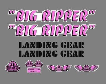 Puede incluir: Un conjunto de pegatinas con el texto "BIG RIPPER" en rosa con bordes negros, "LANDING GEAR" en negro, y varios logotipos de SE Racing en rosa y negro, incluyendo diseños alados y una etiqueta 6061-T6 ALLOY sobre un fondo gris.