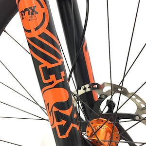 Puede incluir: Horquilla de bicicleta de montaña negra con marca FOX naranja y una calcomanía negra y naranja. La horquilla está unida a una rueda negra con radios naranjas y un rotor de freno negro.