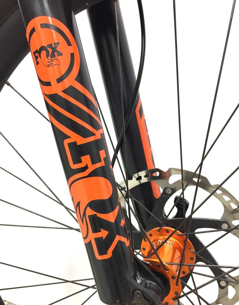 May include: Black mountain bike fork with orange FOX branding and a black and orange decal. The fork is attached to a black wheel with orange spokes and a black brake rotor.