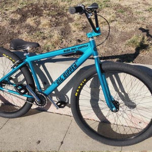 May include: A teal-colored BMX bike with black tires and a black seat. The bike has the text "RIDE OUT SUPPLY" on the frame and "BIG RIPPER" on the side. The tires have the brand name "MAXXIS" in yellow.