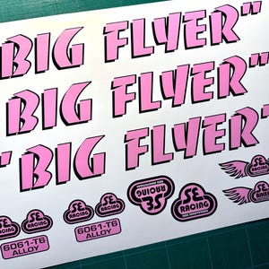 May include: A white decal sheet featuring three sets of the text BIG FLYER in a bold pink font with black outlines. The sheet includes several smaller pink and black logo stickers with wings and the text SE RACING, SE BIKES, and 6061-T6 ALLOY.