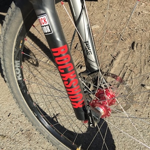 May include: Close-up of a black mountain bike fork with a red "RockShox Pike" logo. The fork is attached to a black wheel with silver spokes and a red hub.