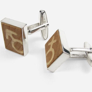 Könnte beinhalten: Ein Paar silberne Manschettenknöpfe mit einer quadratischen Holzeinlage, die ein stilisiertes Symbol zeigt. Das Holz ist hellbraun.