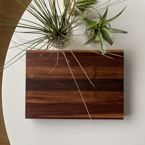 Puede incluir: Una tabla de cortar de madera marrón oscuro con un patrón de veta de madera natural. La tabla está sobre una superficie blanca con tres pequeñas plantas en maceta.