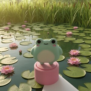 May include: A light mint green plush frog figurine with a white belly and pink cheek accents, perched atop a solid cylindrical pink base. The frog has a simple black line for a mouth and large black eyes, presented as a decorative object.