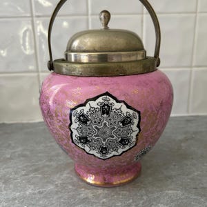 May include: A pink ceramic jar with a silver lid and handle. The jar has a decorative black and white design and gold accents. The handle is arched, and the lid has a small knob. The jar is likely for storage or display.