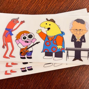 May include: A set of four stickers featuring cartoon characters. The characters are a tall red stick figure with a blue scarf, a pink character with large eyes playing a guitar, a yellow character with a blue and red shirt with a target logo, and a man with a white shirt and black suit playing a keyboard.