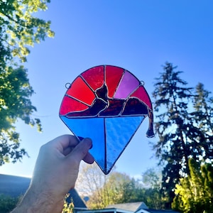 May include: Stained glass cat suncatcher with a geometric design. The cat is depicted in a reclining pose, with a red, pink, and blue color scheme. The suncatcher is held up against a blue sky.
