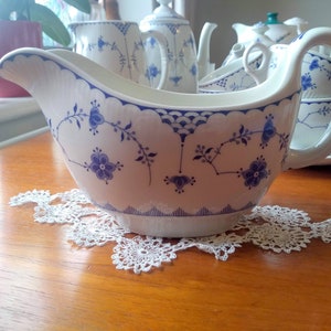 May include: A white gravy boat with a blue floral pattern. The boat has a scalloped edge and a handle. It is sitting on a white doily on a wooden table.