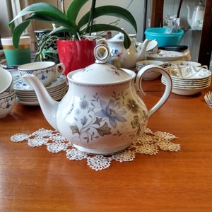 May include: A white teapot with a floral design in shades of blue and green. The teapot has a handle and a spout. It is sitting on a table with a lace doily.