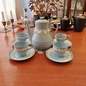 May include: A light blue tea set with white floral detailing. The set includes a teapot, saucers, and teacups. The teapot has a lid and a decorative white floral band. The teacups and saucers have a matching floral design. The set is arranged on a wooden table.