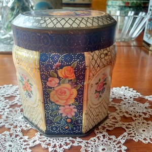 Puede incluir: Una lata hexagonal vintage con un diseño floral azul y dorado. La lata tiene una tapa dorada con un patrón de diamante y está decorada con una variedad de flores, incluyendo rosas, margaritas y otros motivos florales.