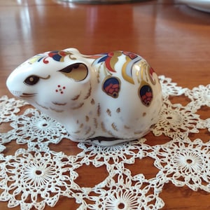 Puede incluir: Una figurita de ratón de porcelana blanca con intrincados detalles pintados en oro, rojo y azul. El ratón está sentado sobre un encaje de ganchillo blanco.