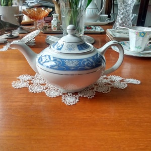 Puede incluir: Una tetera blanca con un diseño floral azul y dorado. La tetera tiene una tapa con un pomo en la parte superior. La tetera está sentada sobre un encaje blanco en una mesa de madera.