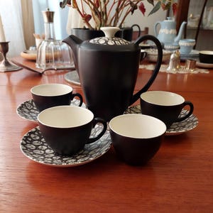 Puede incluir: Juego de té de cerámica negra con interiores blancos, que incluye una tetera y cuatro tazas sobre platillos a juego. Los platillos tienen un patrón en blanco y negro. La tetera tiene una tapa con estampado en blanco y negro.