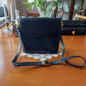 May include: A black leather shoulder bag with a gold buckle and a long adjustable strap. The bag has a flap closure and a small pocket on the front.