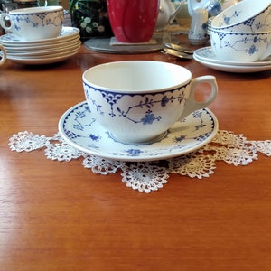 Puede incluir: Taza y platillo de porcelana blanca con diseños florales azules. La taza descansa sobre un platillo a juego, colocado sobre un tapete de encaje blanco. Se ven más tazas y platillos, lo que sugiere un servicio de té.