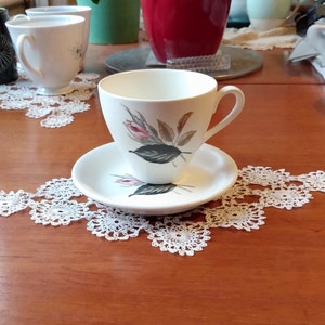 Könnte beinhalten: Eine weiße Teetasse und Untertasse mit Blumenmuster. Die Tasse hat ein zartes Blumenmuster mit rosa und grünen Blättern. Die Untertasse ist schlicht weiß.