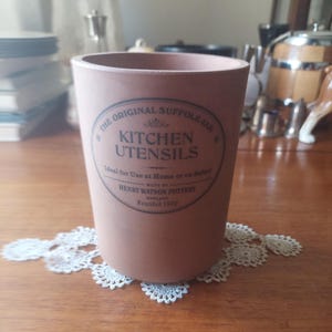 May include: A terracotta-coloured kitchen utensil jar with a printed label reading "THE ORIGINAL SUFFOLK JAR KITCHEN UTENSILS" and "HENRY WATSON POTTERY ENGLAND Founded 1800". It sits on a white lace doily. A kitchenware item.