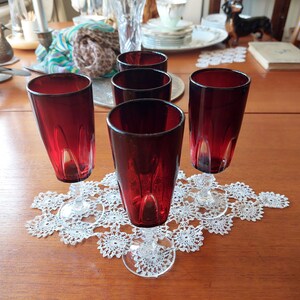 May include: Six red glass champagne flutes with clear stems and bases, arranged on a white crocheted doily on a wooden table.