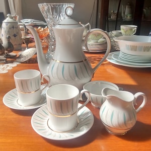 Puede incluir: Un juego de té blanco con un patrón floral verde y dorado. El juego incluye una tetera, una jarra de crema, un azucarero y dos tazas de té con platillos.