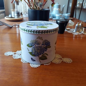 May include: A white, round tin with a lid featuring blue hydrangea flowers and green leaves. The tin is decorated with butterflies and dragonflies. It sits on a white, lace doily on a wooden table. A blue teapot and cups are in the background.