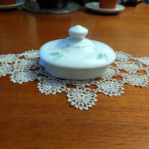Könnte beinhalten: Ein weißer Keramikdeckel mit kleinem Griff, verziert mit grünen floralen Akzenten, liegt auf einer zarten, cremefarbenen Spitzendecke. Der Deckel ist oval und liegt auf einem Holztisch. Die Tischdecke hat ein Blumenmuster.