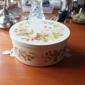 May include: Round, vintage tin box with a cream-colored base and lid. The lid and sides feature a botanical design with berries, leaves, and wheat stalks in shades of brown, green, and red. The box is sitting on a white lace doily.
