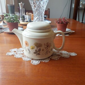 Può includere: Teiera in ceramica color crema con coperchio abbinato, decorata con motivi floreali gialli, arancioni e marroni. La teiera è appoggiata su un centrino di pizzo bianco su un tavolo di legno. Un classico articolo da cucina.