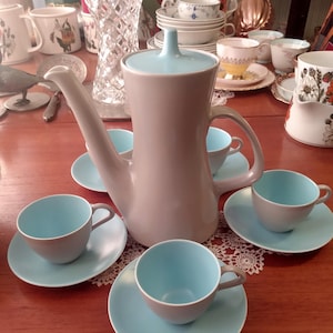 Puede incluir: Una cafetera de cerámica gris y azul con tapa y un juego a juego de cuatro tazas y platillos. Las tazas y los platillos tienen un interior azul claro y un exterior gris.