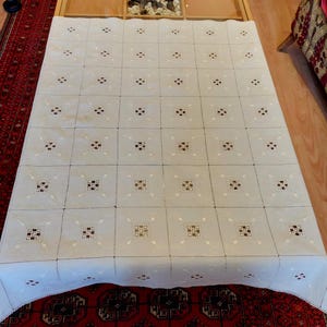 Pode incluir: Uma toalha de mesa creme com um padrão quadrado repetitivo. Cada quadrado apresenta um design central com detalhes escuros e claros. A toalha de mesa está sobre uma superfície de madeira, com um tapete vermelho estampado visível por baixo.