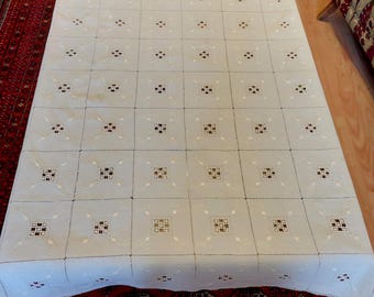 Puede incluir: Un mantel crema con un patrón cuadrado repetitivo. Cada cuadrado presenta un diseño central con acentos oscuros y claros. El mantel está sobre una superficie de madera, con una alfombra roja estampada visible debajo.