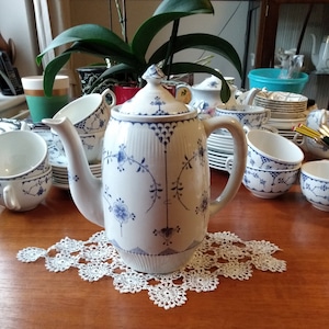 Puede incluir: Una cafetera de cerámica blanca con un patrón floral azul y blanco. La cafetera tiene una tapa y un pico. Está sentada sobre un encaje blanco en una mesa de madera.