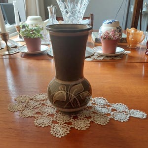 Può includere: Un vaso in ceramica marrone con un disegno a foglie. Il vaso ha un collo stretto e una base larga. È appoggiato su un centrino di pizzo bianco.