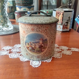 Lata de galletas vintage. Hermosas escenas de pueblos de montaña y lago en paneles redondos sobre un fondo floral estilizado en crema, dorado y naranja.