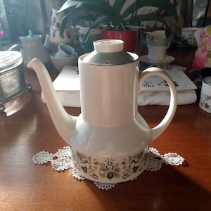 Könnte beinhalten: Eine weiße Teekanne aus Keramik mit einem grünen Deckel und einem Blumenmuster. Die Teekanne steht auf einer weißen Spitze auf einem Holztisch.