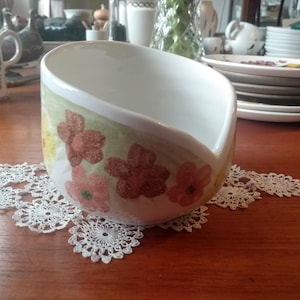 Könnte beinhalten: Weißer Keramiknapf mit einem Blumendesign in Rot- und Gelbtönen. Die Schale steht auf einer weißen Spitzendecke. Der Hintergrund umfasst einen Holztisch und anderes Geschirr.
