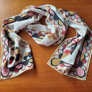 May include: A long rectangular scarf featuring a white background with a repeating scalloped pattern in shades of pink, black, gold, and light blue. The edges are finished with a thin gold border, and the fabric displays a smooth, soft texture.